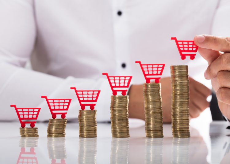 Businessperson's hand placing red shopping cart over increasing stacked coins