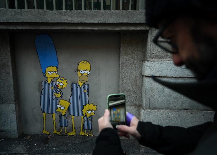 A man takes a photo of a mural by Italian street artist AleXsandro Palombo entitled "Track 21 The Simpsons deported to Auschwitz" on January 26, 2023 near Milan's Central railway station, on the occasion of the upcoming International Holocaust Remembrance Day. - "The first work is a portrait of the family before deportation, in the second the now imprisoned Simpsons appear emaciated, skeletal and wearing the striped uniforms of the concentration camps, deprived of dignity" the artist stated. (Photo by Piero CRUCIATTI / AFP) / RESTRICTED TO EDITORIAL USE - MANDATORY MENTION OF THE ARTIST UPON PUBLICATION - TO ILLUSTRATE THE EVENT AS SPECIFIED IN THE CAPTION